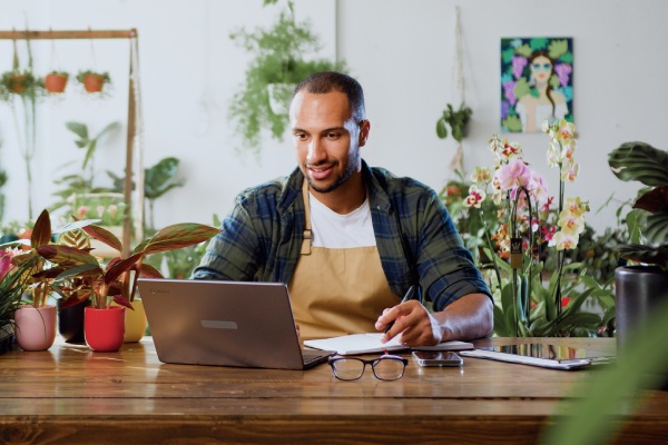 Man florist using laptop, typing and taking inventory in Plant Store. Botanist working, small business owner. Florist working on computer at workplace.