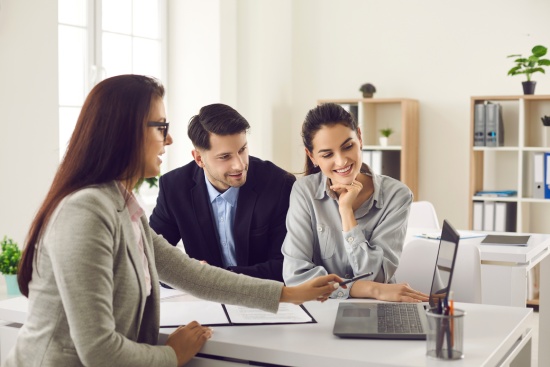 Happy future house buyers meeting real estate agent. Professional realtor talking to clients and offering flats options on computer. Smiling couple consulting bank worker or loan broker at her office