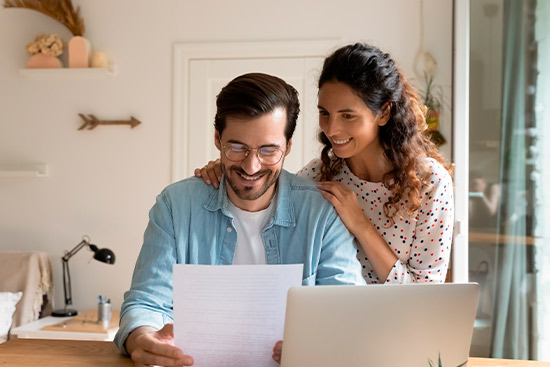 couple smiling reading a paper in front a computer. 